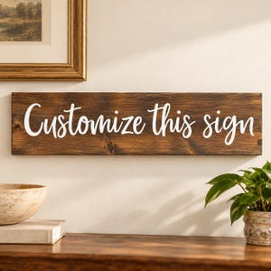 May include: A rectangular wooden sign with a dark brown stained finish and visible wood grain. The text "Customize this sign" is written across the center in a white, cursive script font. The sign is mounted horizontally on a plain wall.
