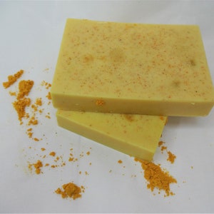 Puede incluir: Dos barras de jab&oacute;n amarillo con textura granulada, sobre una superficie blanca con polvo amarillo esparcido a su alrededor.