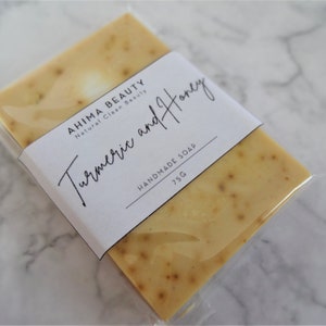 Puede incluir: Una barra de jab&oacute;n artesanal de c&uacute;rcuma y miel envuelta en pl&aacute;stico transparente. La etiqueta dice "AHIMA BEAUTY Natural Clean Beauty C&uacute;rcuma y Miel JAB&Oacute;N HECHO A MANO 75G".