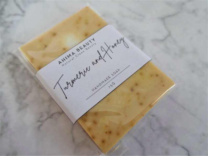 Puede incluir: Una barra de jab&oacute;n artesanal de c&uacute;rcuma y miel envuelta en pl&aacute;stico transparente. La etiqueta dice "AHIMA BEAUTY Natural Clean Beauty C&uacute;rcuma y Miel JAB&Oacute;N HECHO A MANO 75G".