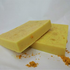 Puede incluir: Dos barras de jab&oacute;n amarillo con motas de color naranja. El jab&oacute;n est&aacute; sobre una superficie blanca con un poco de polvo naranja esparcido alrededor.