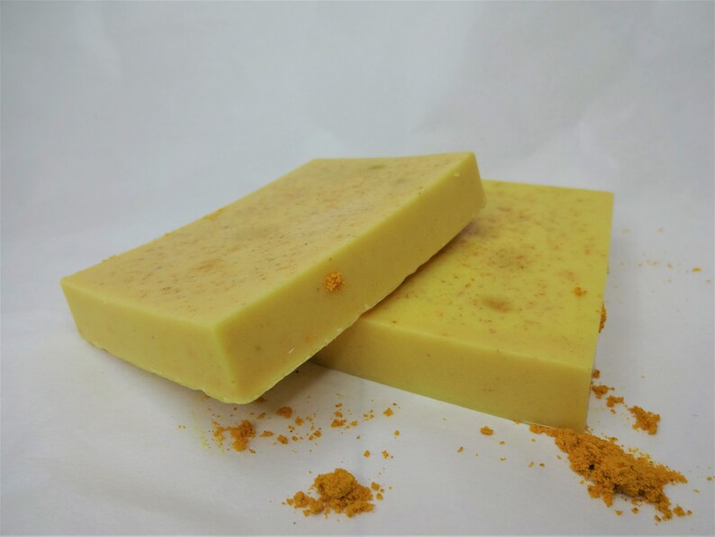 Puede incluir: Dos barras de jab&oacute;n amarillo con motas de color naranja. El jab&oacute;n est&aacute; sobre una superficie blanca con un poco de polvo naranja esparcido alrededor.