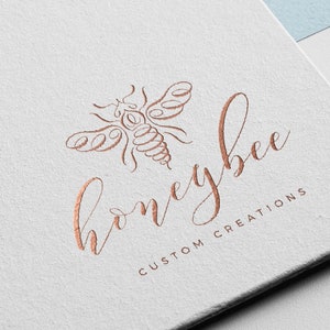 Può includere: Un logo per un'azienda chiamata "honeybee custom creations" con un disegno stilizzato di un'ape in color rame.