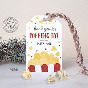 May include: A popcorn-themed gift tag with the text "Thank you for POPPING BY!" in red and black. The tag features a popcorn graphic and is tied with a metallic cord. Scattered popcorn kernels are at the base.
