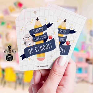 Puede incluir: Dos etiquetas de regalo blancas con un dise&ntilde;o de l&aacute;piz y b&uacute;ho. Las etiquetas dicen "Happy First Day of School!" en azul marino sobre un l&aacute;piz amarillo. Tambi&eacute;n aparece una manzana roja. Las etiquetas se sostienen en una mano.