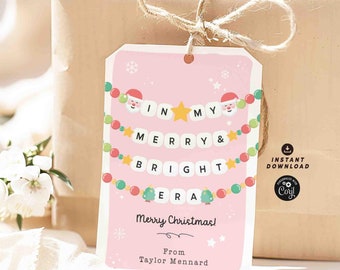 Könnte beinhalten: Rosafarbener Weihnachtsgeschenkanhänger mit einer festlichen Girlande aus Perlen und Buchstaben, die "IN MY MERRY & BRIGHT ERA" buchstabieren. Der Anhänger zeigt eine Illustration von Weihnachtsmann und den Text "Frohe Weihnachten! Von Taylor Mennard".