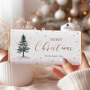 Puede incluir: Un envoltorio de barra de chocolate blanco con una ilustración de acuarela de un pino y estrellas doradas brillantes. El texto "Merry Christmas" está escrito en cursiva dorada. El texto "The Gail Family - 202X" está escrito en dorado debajo del saludo. La parte inferior del envoltorio tiene el texto "cutepartydash.com".