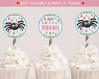 以下が含まれることがあります： 黒のクモにピンクのリボン、ターコイズとゴールドのグリッター、そして「INDIANA」の文字が書かれた、編集可能なカップケーキのトッピング。トッピングは、白いフロスティングをかけた白いカップケーキの上に載っています。