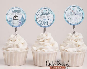 Puede incluir: Tres adornos para cupcakes con temas de invierno. El primer adorno presenta un pingüino con el texto "Winter One-derland". El segundo adorno presenta copos de nieve y el texto "Snow Much Fun! To Be One". El tercer adorno presenta un oso polar y un pingüino con una escena de montaña en el fondo.
