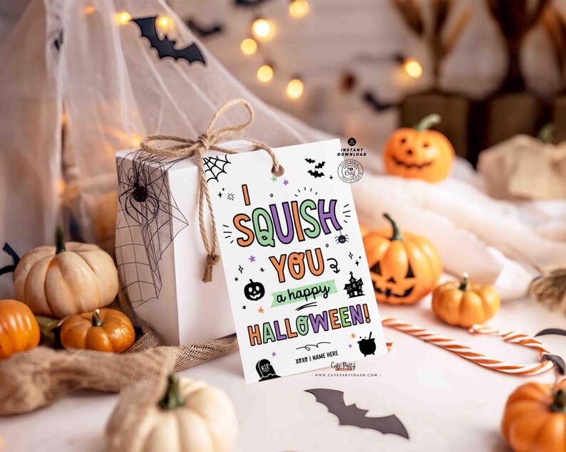 May include: A white gift box tied with twine, adorned with a Halloween-themed tag that reads "I squish you a happy Halloween!" surrounded by pumpkins, bats, and a spiderweb. The scene is festive with decorative lights.