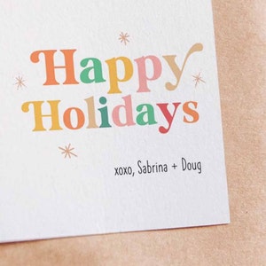May include: A white card with the text "Happy Holidays" in colorful letters. The text "xoxo, Sabrina + Doug" is printed below the greeting.