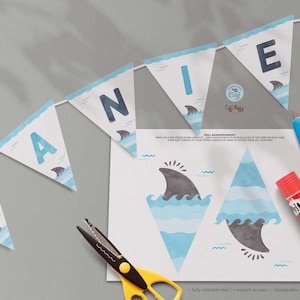 May include: A printable shark-themed banner with the letters "DANIEL" in blue on a white background. The banner is decorated with blue and white waves and grey shark fins. The image also includes instructions for assembling the banner, a pair of scissors, and a glue stick.