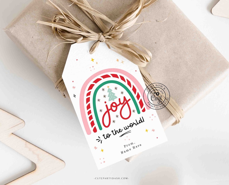 May include: A white gift tag with a colorful rainbow design and the text "joy to the world!" in red. The rainbow is made of pink, green, and red stripes. There are small white snowflakes and stars scattered around the rainbow. The text "From, Name Here" is at the bottom of the tag.