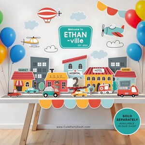 May include: A birthday party decoration set with a city theme. The set includes a banner that says "Welcome to Ethan-ville Est. 2022", a cake topper with the number 2, and various buildings, vehicles, and balloons. The colors are bright and cheerful, with red, blue, yellow, and green being prominent.