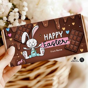 Può includere: Una barretta di cioccolato con un involucro marrone raffigurante un coniglietto dei cartoni animati e le parole "HAPPY Easter" in rosa. L'involucro include anche quadratini di cioccolato, cuori e la scritta "from Name".