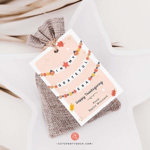 K&ouml;nnte beinhalten: Ein rosa-wei&szlig;es Geschenkanh&auml;nger mit einer Girlande aus Herbstbl&auml;ttern und dem Text "In my grateful heart" und "Happy Thanksgiving" mit einem handschriftlichen "From Taylor Maynard".