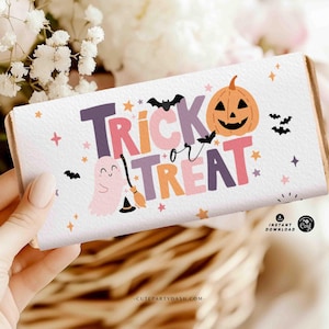 May include: A white candy bar wrapper with the words "Trick or Treat" in purple, orange, and pink. The design includes a smiling jack-o'-lantern, bats, stars, and a ghost with a witch's broom. The wrapper is held by a person.
