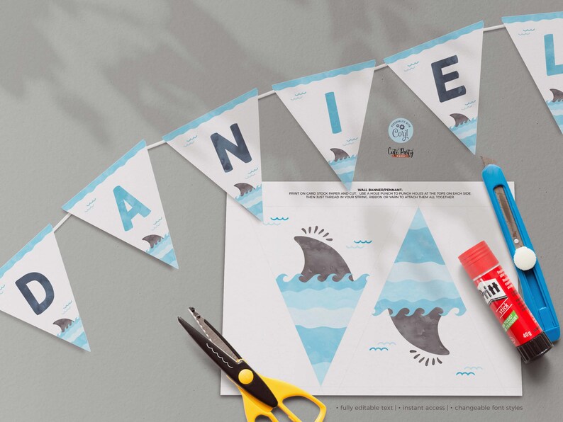 May include: A printable shark-themed banner with the letters "DANIEL" in blue on a white background. The banner is decorated with blue and white waves and gray shark fins. The image also includes instructions for assembling the banner, a pair of scissors, and a glue stick.