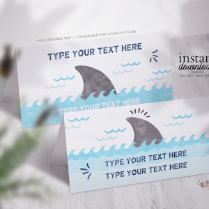 May include: Editable printable place cards featuring a shark fin and blue watercolour waves. The text "Type your text here" is displayed on the cards. The cards are 4x2 inches when folded.