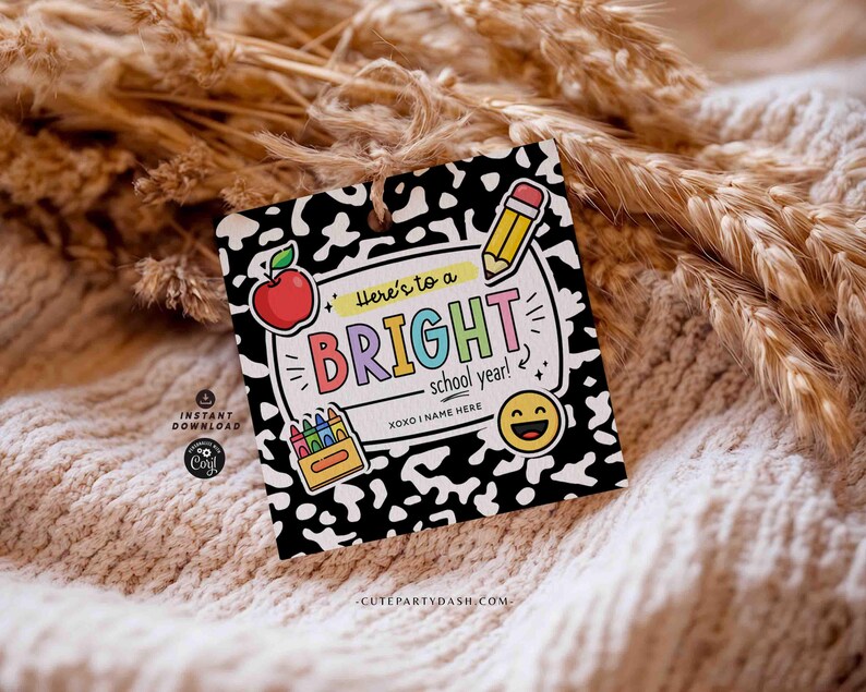 Op de afbeelding: Een vierkant label met een zwart-wit patroon, met een appel, potlood, kleurpotloden en een smiley. Het label leest "Here's to a BRIGHT school year!" in kleurrijke letters. Het label is vastgebonden met een touwtje.