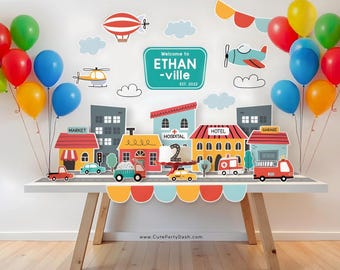 May include: A birthday party table setting with a city theme. The table is decorated with paper cutouts of buildings, cars, and a helicopter. There are balloons in red, yellow, blue, and green. The cake is decorated with a number 2 and a red cross. The banner says "Welcome to Ethan-ville Est. 2022".