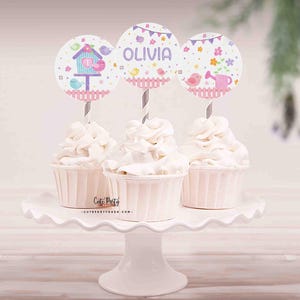 May include: Three cupcakes decorated with pink and blue birdhouse themed toppers. The toppers have the name "Olivia" on one of them.