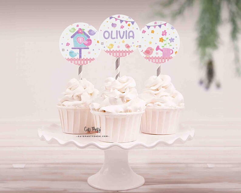 May include: Three cupcakes decorated with pink and blue birdhouse themed toppers. The toppers have the name "Olivia" on one of them.