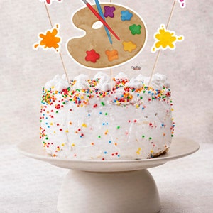 Pu&ograve; includere: Una torta bianca decorata con codette colorate e sormontata da decorazioni per torte a forma di tavolozza e schizzi di vernice. La torta &egrave; su un supporto per torte bianco.