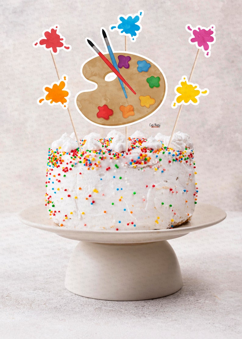 Pu&ograve; includere: Una torta bianca decorata con codette colorate e sormontata da decorazioni per torte a forma di tavolozza e schizzi di vernice. La torta &egrave; su un supporto per torte bianco.