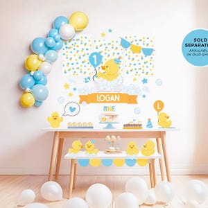 May include: A yellow and blue birthday party backdrop with a rubber ducky theme. The backdrop features a yellow rubber ducky with a party hat, a blue balloon, and the number one. The backdrop also includes the name "Logan" and the word "One".