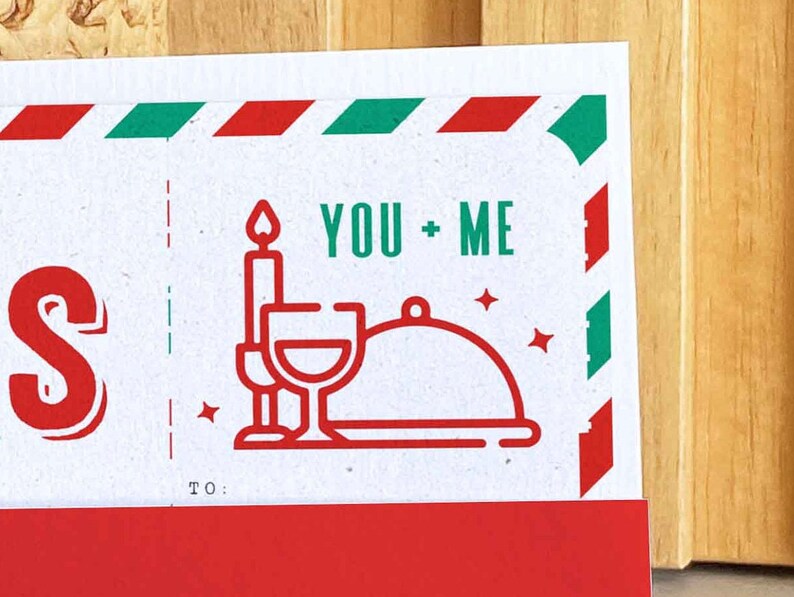 May include: A white card with red and green accents. The card features the text "YOU + ME" in green, along with illustrations of a candle, wine glasses, and a covered dish. The word "S" is in red, and the bottom has a red border.