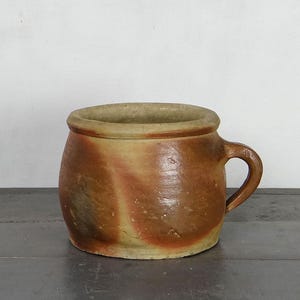 May include: A rustic, handmade ceramic mug with a rounded body and a small handle. The mug features a mottled brown and tan color scheme, with visible texture and imperfections, suggesting an aged appearance. The rim is a lighter tan color.