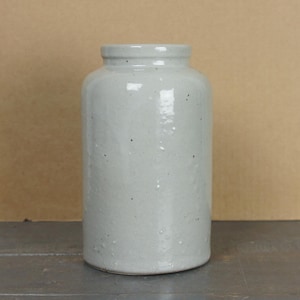 Puede incluir: Un tarro de gres gris claro con un cuerpo cilíndrico y un borde ligeramente ensanchado. El tarro tiene un aspecto moteado y un acabado liso y brillante. El tarro es una pieza vintage, probablemente utilizada para el almacenamiento.