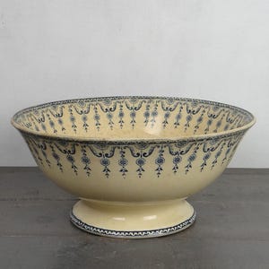 May include: A cream-colored ceramic bowl with a pedestal base, featuring a dark blue decorative pattern of hanging floral garlands and swags around the interior and exterior rim, and a matching patterned border on the base.