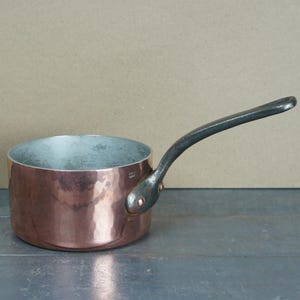 May include: A copper saucepan with a long, dark handle. The pot has a hammered finish and a silver-coloured interior. The handle is curved and attached to the side of the pot. The saucepan is on a grey surface.