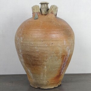May include: A large, bulbous earthenware jug with a textured, tan and reddish-brown glazed surface. The vessel features a narrow central neck, a small circular opening, and two thick, ribbed handles attached to the upper shoulder area.