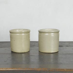 May include: Two small, cylindrical, stoneware jars with a light beige glaze. The jars have a rounded rim and a slightly tapered body. They are displayed on a dark wooden surface against a white background. These jars are suitable for kitchen or home decor.