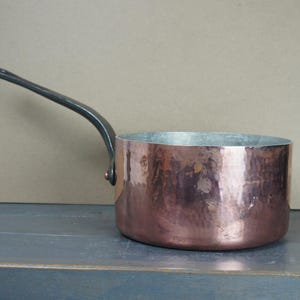 Può includere: Una casseruola in rame con finitura martellata e manico in metallo scuro. La pentola ha una forma arrotondata e un interno color argento. Il manico è curvo verso l'alto. La padella è progettata per cucinare.