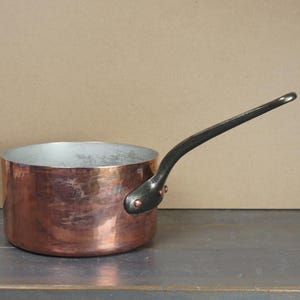May include: A copper saucepan with a silver interior and a long, black handle. The pot is cylindrical and sits on a dark surface, with a neutral background.