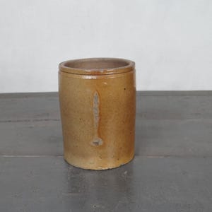 Peut inclure: Un pot cylindrique en grès antique avec un glaçage brun clair. Le pot a une forme légèrement conique et un bord arrondi. Il est probablement utilisé pour le stockage et a un aspect rustique et vieilli.