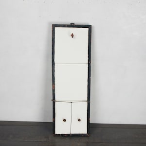 May include: A rectangular, vintage-style wall hanging with a black frame and a white interior. The frame has distressed red accents. A wire is strung across the back for hanging. Two small wooden pegs are visible on the white surface.