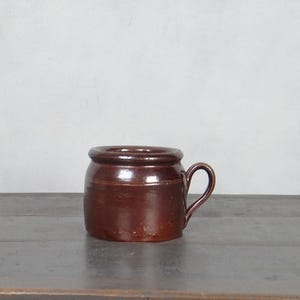 Könnte beinhalten: Eine kleine, braune Keramik-Tasse mit Henkel. Die Tasse hat einen abgerundeten Körper und einen leicht ausgestellten Rand. Der Henkel ist gebogen und an der Seite der Tasse befestigt. Die Tasse steht auf einer Holzoberfläche.