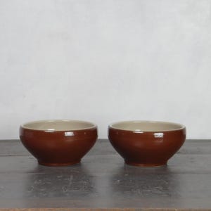 Può includere: Due piccole ciotole rotonde in ceramica. L'esterno è di un marrone ricco e lucido, mentre l'interno è beige chiaro. Le ciotole sono poste fianco a fianco su una superficie di legno scuro, contro uno sfondo bianco.