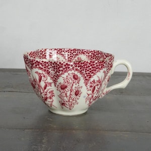 May include: A white teacup with a red floral pattern. The cup has a scalloped rim and a handle.