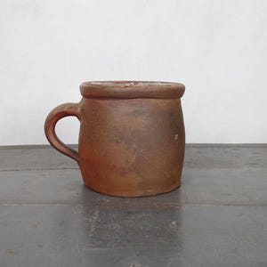 May include: A rustic, handmade brown ceramic mug with a curved handle. The mug has a wide, slightly flared rim and a rounded body. The mug is unglazed and has a textured surface, perfect for coffee or tea.