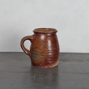May include: A rustic, handmade ceramic mug with a handle. The mug is primarily brown with reddish and tan hues, and a slightly lighter rim. The mug sits on a dark gray surface against a white background. The mug appears to be hand-thrown.
