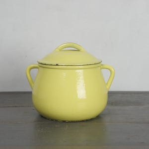 Puede incluir: Una olla de esmalte amarilla con tapa. La olla tiene dos asas y un asa pequeña en la tapa. La olla es de color amarillo claro con un borde negro alrededor de la tapa. La olla está sobre una superficie gris.