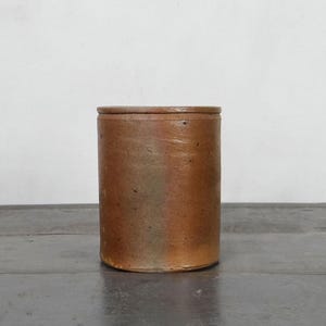 Könnte beinhalten: Ein zylindrischer, antiker Steinguttopf mit Deckel. Der Topf hat eine warme braune Farbe mit subtilen Variationen und einer leicht rauen Textur. Schlichtes, rustikales Design, geeignet für Küche oder Wohnkultur.