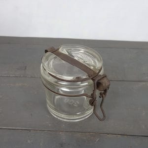 Puede incluir: Un tarro de vidrio transparente con tapa de metal y cierre de alambre. El tarro tiene una superficie texturizada y está vacío.