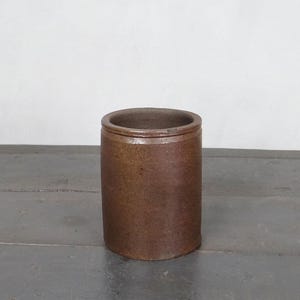 Peut inclure: Un pot en grès marron de forme cylindrique avec un bord légèrement évasé. Le pot antique a une apparence rustique et vieillie, suggérant qu'il pourrait être vintage ou une reproduction. La surface présente de subtiles variations de couleur et de texture.